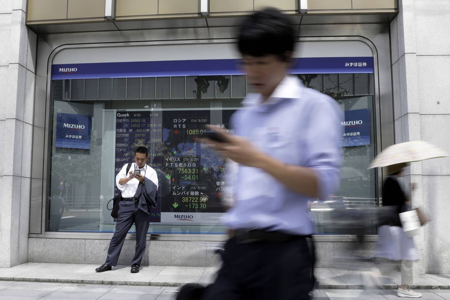 Un peatón que utiliza su teléfono inteligente se encuentra frente a un tablero electrónico de valores fuera de una firma de valores en Tokio, Japón, el jueves 30 de agosto de 2018. Fotógrafo: Kiyoshi Ota/Bloomberg