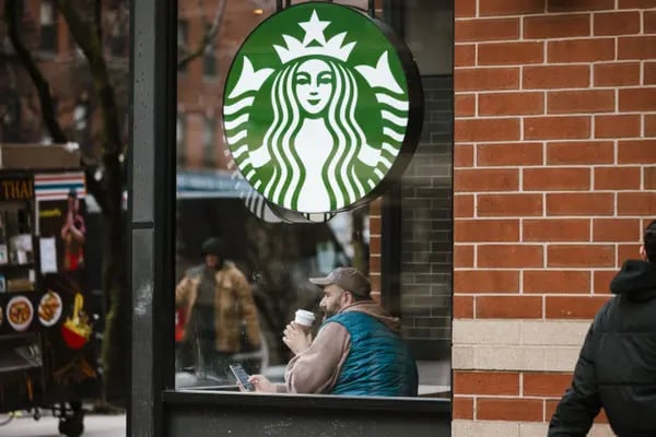 Fachada de uma loja da Starbucks. É possível ver o logotipo, um círculo verde com um desenho de sereia com duas caudas. Dentro da loja está um cliente sentado à uma mesa e tomando um copo de café. Ele aprecia a vista.