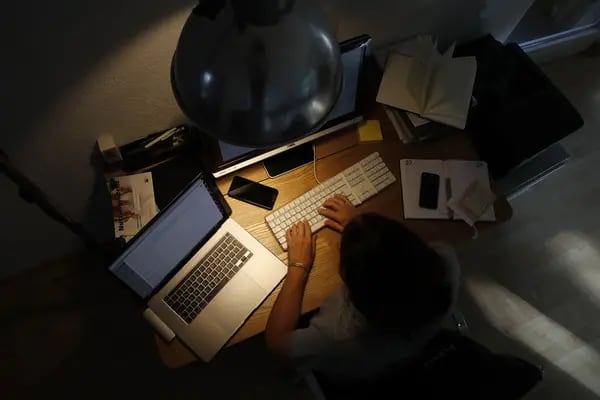 Foto de ângulo superior mostrando uma escrivaninha iluminada por uma luminária de luz amarela. Tem uma pessoa trabalhando em um computador e rodeada por um laptop, um celular e outros itens de trabalho
