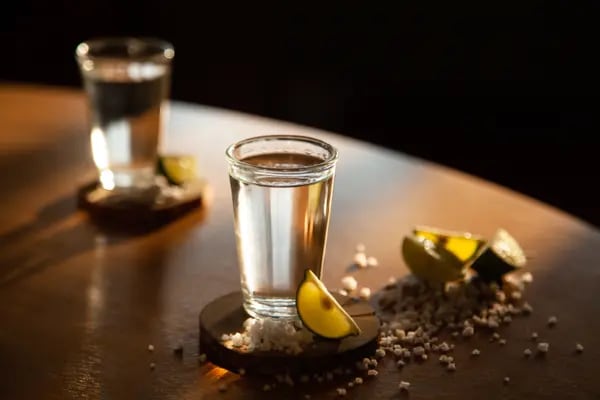 Dos vasos con tequila acompañados de limones y sal de grano sobre una mesa de madera (Foto: Pexels).