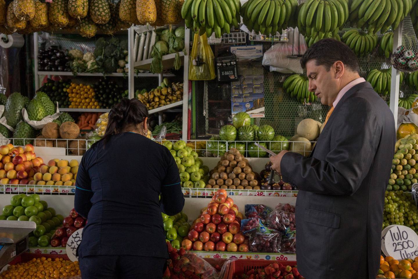 Un hombre revisa su lista de compras mientras un trabajador selecciona frutas para él en un mercado de frutas