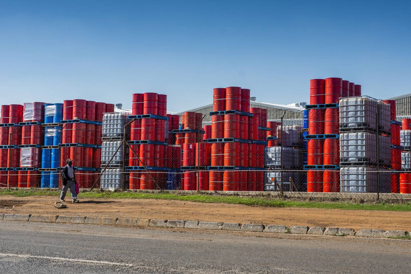 A pedestrian wearing a protective face mask walks by oil barrels stacked beyond security fencing at a facility in the Alrode district of Johannesburg, South Africa, on Tuesday, April 21, 2020. The oil meltdown accelerated, with huge losses sweeping through markets as the world runs out of places to store unwanted crude and grapples with negative pricing. Photographer: Waldo Swiegers/Bloomberg