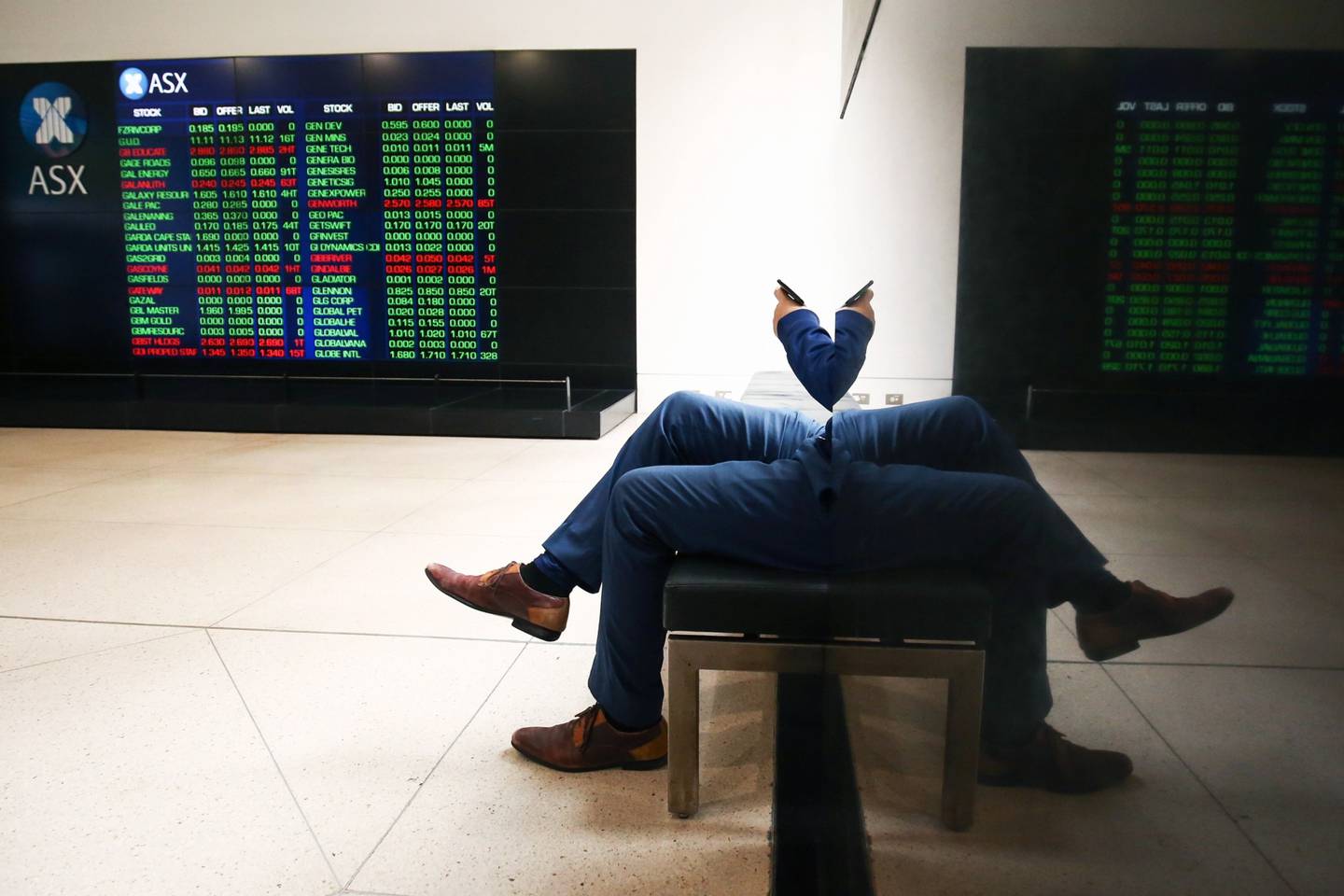 Un hombre que mira su teléfono se refleja en una pared mientras un tablero electrónico muestra información bursátil en la Bolsa de Valores de Australia, operada por ASX Ltd., en Sídney, Australia, el lunes 20 de mayo de 2019.