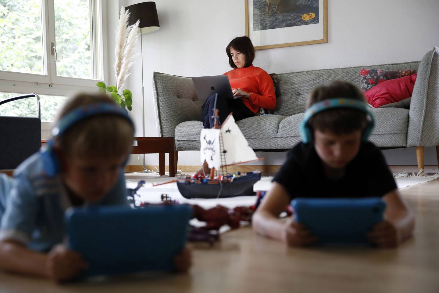 Una mujer trabaja en una computadora portátil de Apple Inc. en el sofá mientras los niños utilizan dispositivos de tableta, en Berna, Suiza, el miércoles 19 de agosto de 2020.