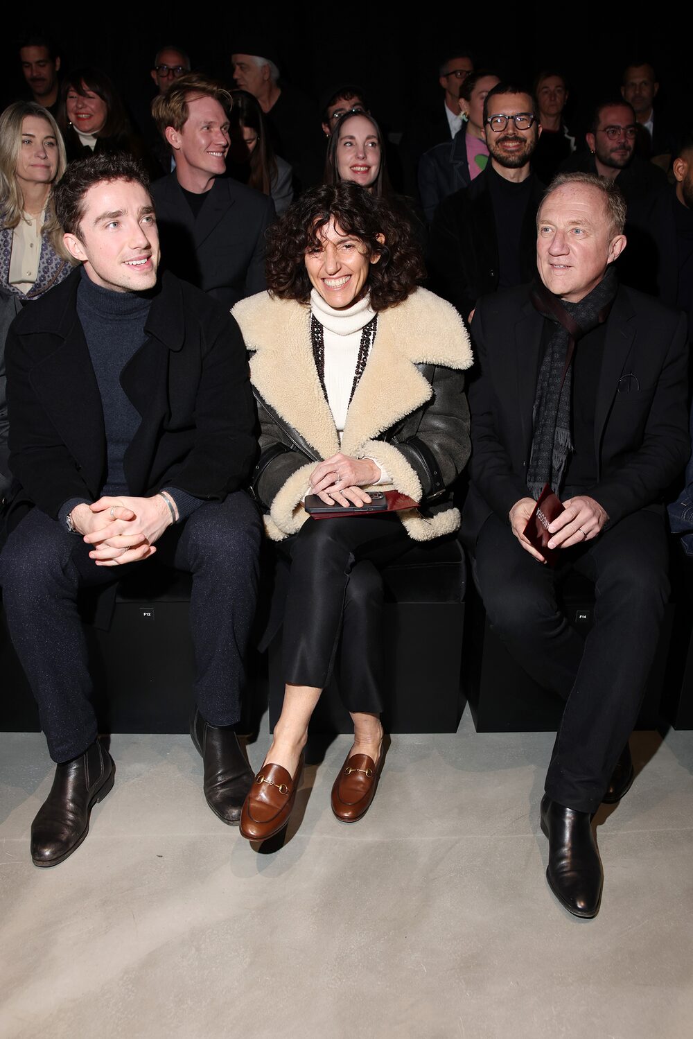 Três pessoas em foco. Elas estão na primeira fila de um desfile de moda. Nas pontas, dois homens e no meio, uma mulher. Três pessoas em foco. Elas estão na primeira fila de um desfile de moda. Nas pontas, dois homens e no meio, uma mulher.