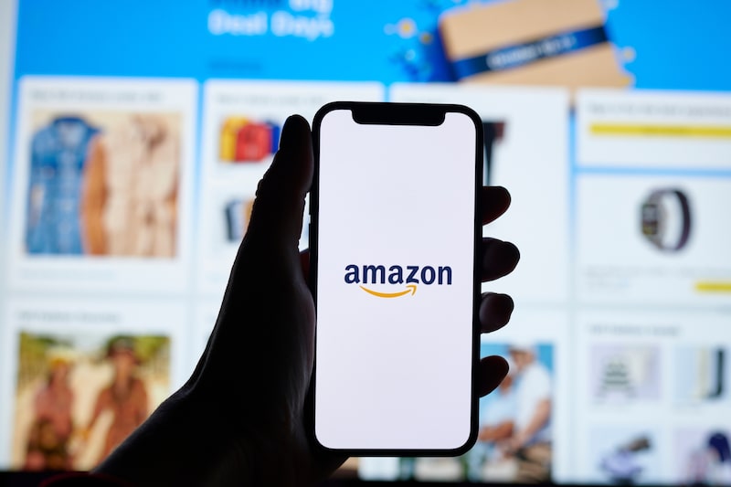 Una persona sostiene un teléfono móvil con el logotipo de Amazon, frente a una pantalla que muestra productos de la plataforma de comercio electrónico. Una persona sostiene un teléfono móvil con el logotipo de Amazon, frente a una pantalla que muestra productos de la plataforma de comercio electrónico.