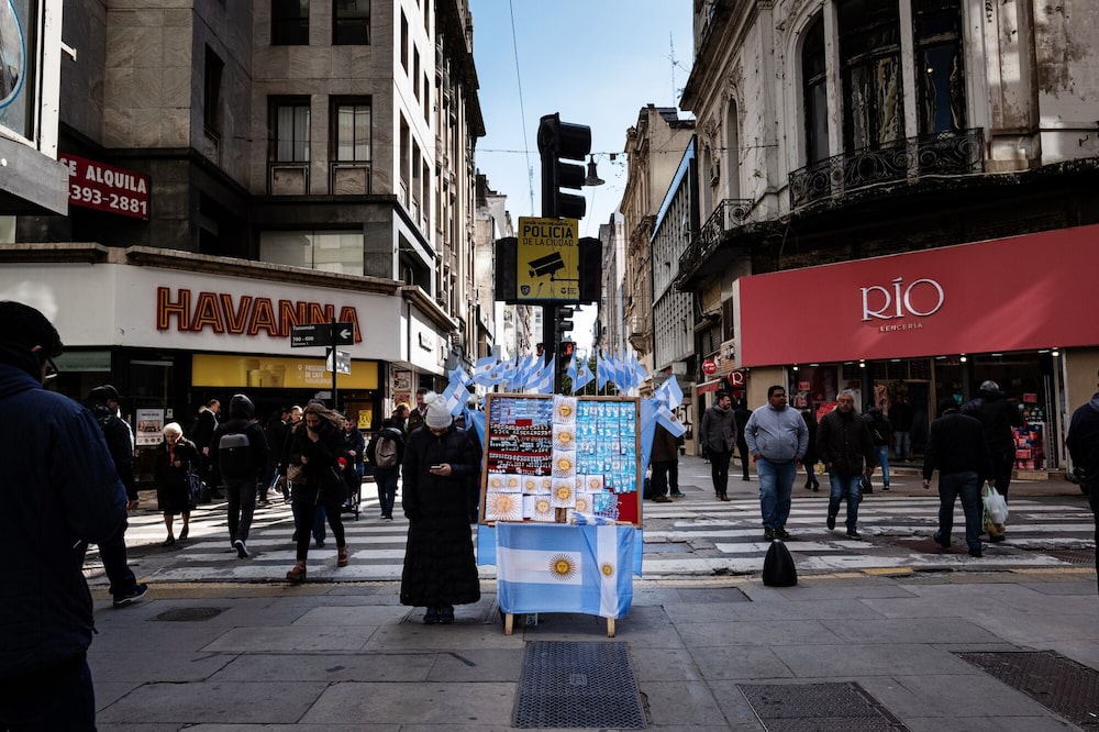 Unos peatones pasan junto a un vendedor ambulante que ofrece artículos con la bandera argentina en una calle de Buenos Aires, Argentina. Unos peatones pasan junto a un vendedor ambulante que ofrece artículos con la bandera argentina en una calle de Buenos Aires, Argentina.