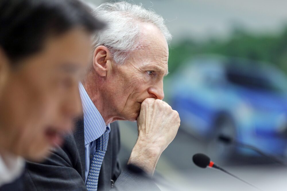 No primeiro plano, um rosto de perfil, não é possível ver suas características físicas por conta do foco, que fica no segundo plano, em um homem grisalho, também de perfil e com olhar consternado No primeiro plano, um rosto de perfil, não é possível ver suas características físicas por conta do foco, que fica no segundo plano, em um homem grisalho, também de perfil e com olhar consternado