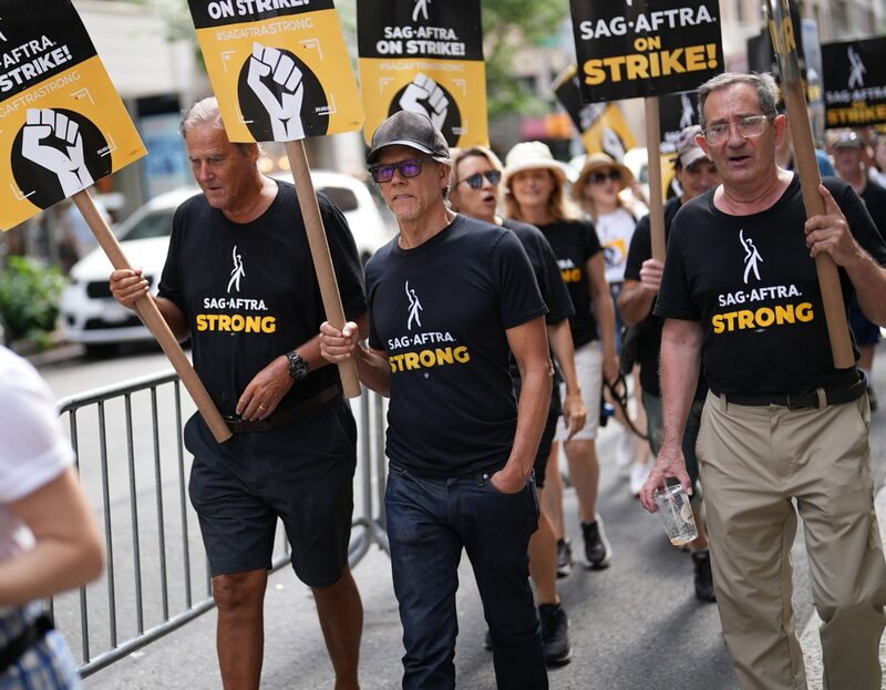 Foto de diversas pessoas, entre elas o ator Kevin Bacon, com camisetas e cartazes relacionados à greve de atores Foto de diversas pessoas, entre elas o ator Kevin Bacon, com camisetas e cartazes relacionados à greve de atores