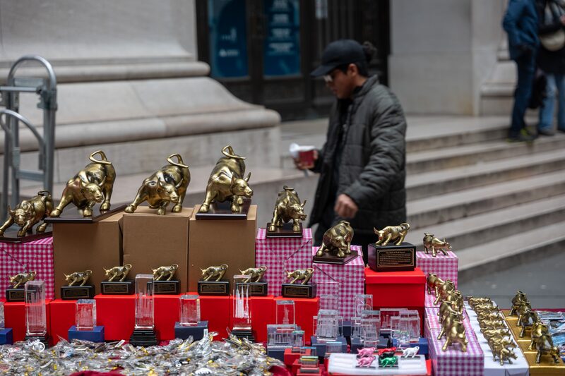 Em primeiro plano, uma mesa repleta de miniaturas de touros dourados em referência ao famoso ponto turístico do Touro em Wall Street. Em segundo plano, um homem de vestes escuras, ele está desfocado Em primeiro plano, uma mesa repleta de miniaturas de touros dourados em referência ao famoso ponto turístico do Touro em Wall Street. Em segundo plano, um homem de vestes escuras, ele está desfocado