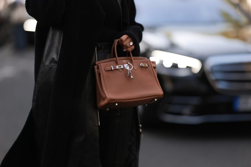 Imagem de uma bolsa Birkin, da Hermes, na cor caramelo, sendo segurada por uma pessoa de gênero não identificado que veste roupas pretas Imagem de uma bolsa Birkin, da Hermes, na cor caramelo, sendo segurada por uma pessoa de gênero não identificado que veste roupas pretas