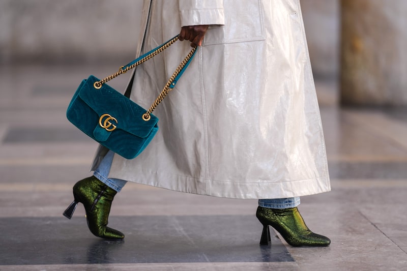 Mulher vista do quadril para baixo, ela está com um casaco branco tapando suas calças e ela usa botas pretas. Na mão, uma bolsa azul-petróleo Mulher vista do quadril para baixo, ela está com um casaco branco tapando suas calças e ela usa botas pretas. Na mão, uma bolsa azul-petróleo