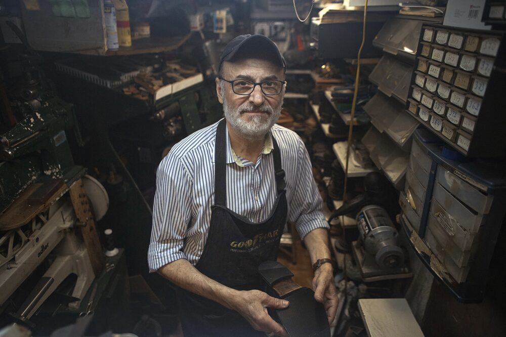 Homem em uma sapataria. Ele veste camisa clara, um avental escuro e um boné também escuro. Tem uma expressão neutra. Usa óculos e tem cabelo e barba brancos Homem em uma sapataria. Ele veste camisa clara, um avental escuro e um boné também escuro. Tem uma expressão neutra. Usa óculos e tem cabelo e barba brancos