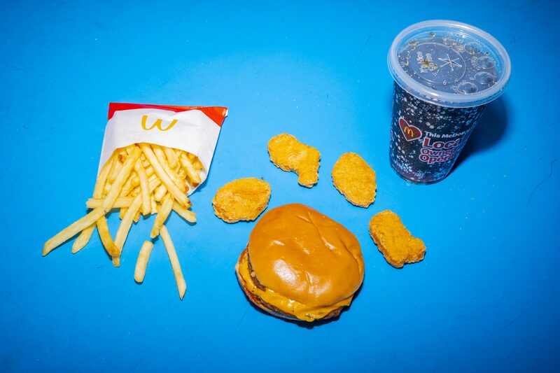 Da esquerda para a direita: um pacote de batatas fritas, um lanche de hambúrguer rodeado por quatro nuggets e um copo de refrigerante de cor escura. O pacote de batatas e o copo de refrigerante levam o logotipo do McDonalds: uma letra M de cor dourada Da esquerda para a direita: um pacote de batatas fritas, um lanche de hambúrguer rodeado por quatro nuggets e um copo de refrigerante de cor escura. O pacote de batatas e o copo de refrigerante levam o logotipo do McDonalds: uma letra M de cor dourada