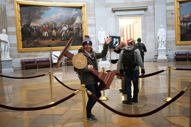 Adam Johnson posando com o púlpito da presidente da Câmara, Nacy Pelosi, durante a invasão do Capitólio Adam Johnson posando com o púlpito da presidente da Câmara, Nacy Pelosi, durante a invasão do Capitólio