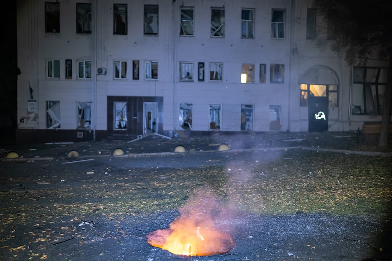 Un edificio con ventanas destruidas y daños visibles tras un ataque, mientras un incendio arde en el exterior. Un edificio con ventanas destruidas y daños visibles tras un ataque, mientras un incendio arde en el exterior.
