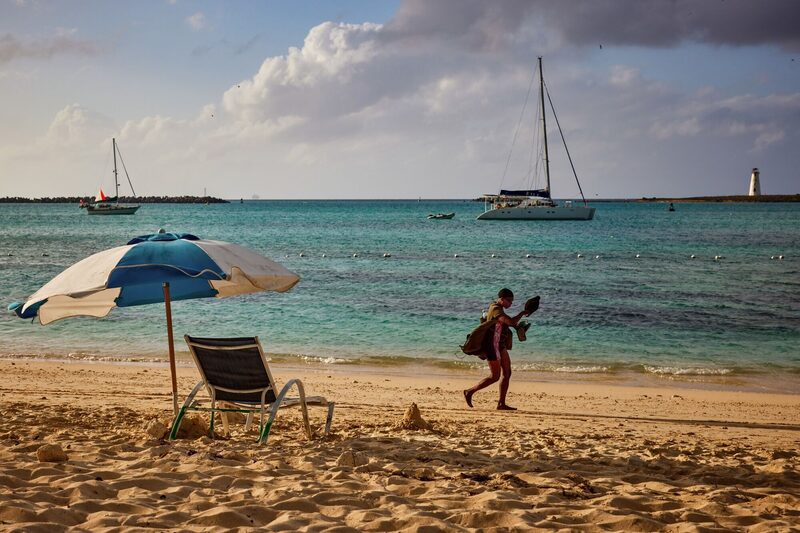 Pessoa passeando em uma praia. É possível ver barcos ao longe e, em primeiro plano, uma cadeira de praia e um guarda-sol Pessoa passeando em uma praia. É possível ver barcos ao longe e, em primeiro plano, uma cadeira de praia e um guarda-sol