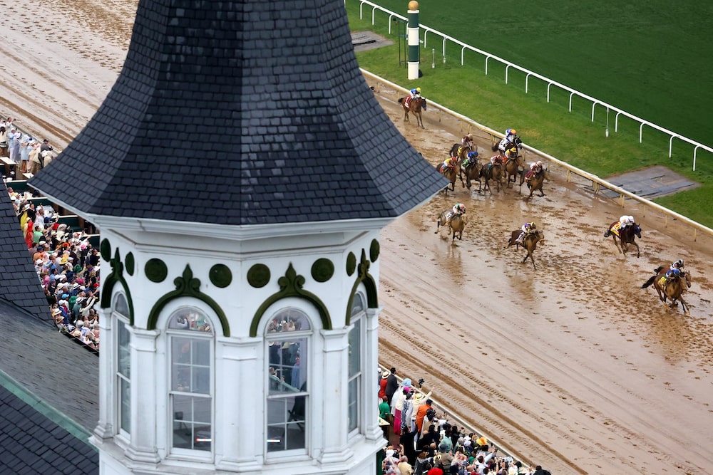 Vista aérea de la recta final del Kentucky Derby, con los caballos avanzando sobre una pista húmeda ante tribunas llenas. Vista aérea de la recta final del Kentucky Derby, con los caballos avanzando sobre una pista húmeda ante tribunas llenas.