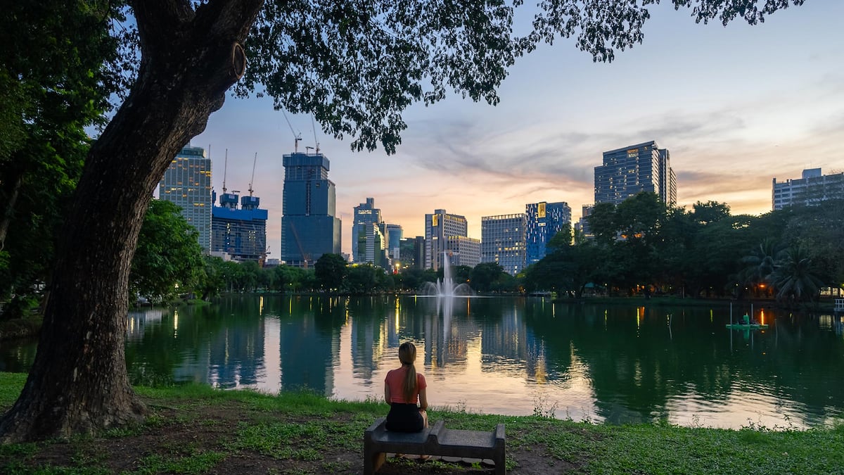 Vista trasera de una mujer sentada en un banco del Parque Lumpini, con el paisaje urbano de Bangkok al atardecer como telón de fondo.