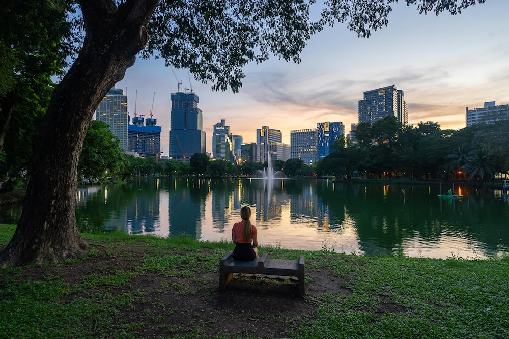 Vista trasera de una mujer sentada en un banco del Parque Lumpini, con el paisaje urbano de Bangkok al atardecer como telón de fondo. Vista trasera de una mujer sentada en un banco del Parque Lumpini, con el paisaje urbano de Bangkok al atardecer como telón de fondo.
