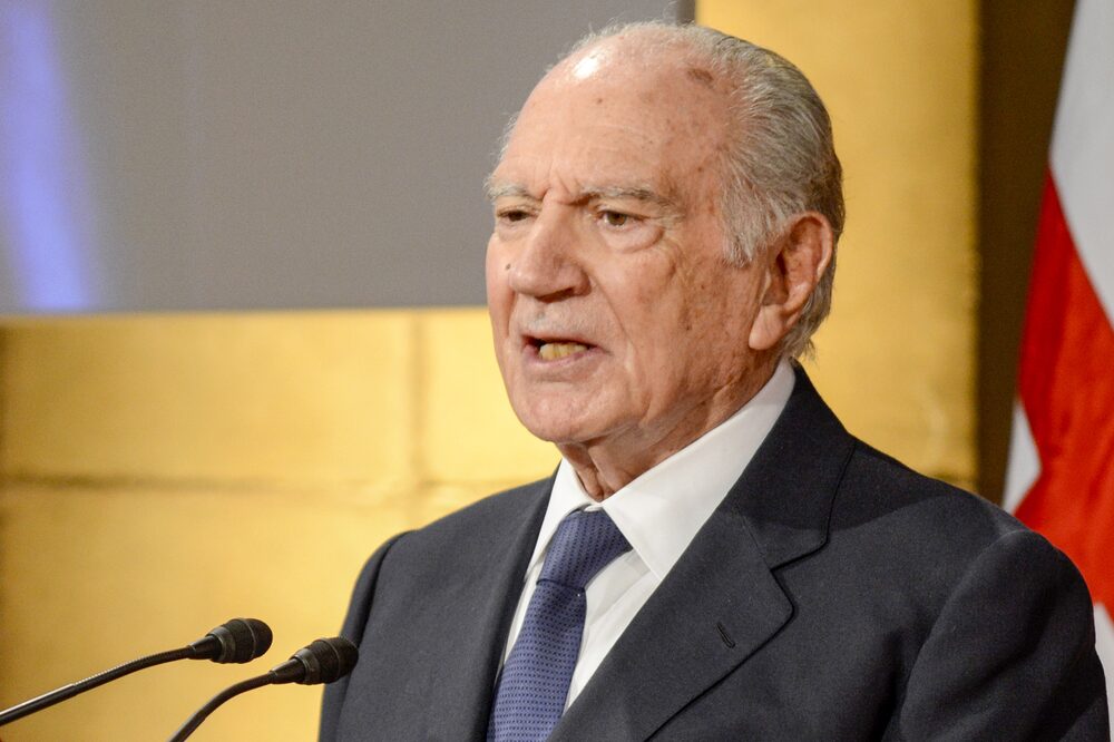 Homem de terno escuro e camisa clara. Ele usa uma gravata azul acinzentado e esta falando em um microfone Homem de terno escuro e camisa clara. Ele usa uma gravata azul acinzentado e esta falando em um microfone