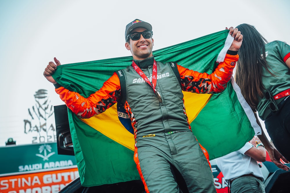 Lucas Moraes com a bandeira do Brasil no Rally Dakar 2026, na Arábia Saudita, com a medalha de competidor no pescoço. Lucas Moraes com a bandeira do Brasil no Rally Dakar 2026, na Arábia Saudita, com a medalha de competidor no pescoço.