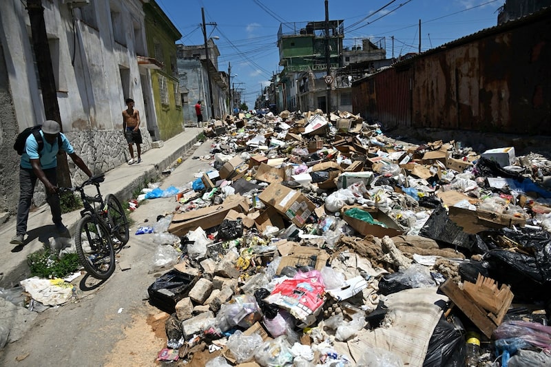 Acumulación de basura bloquea una calle en un barrio urbano de Cuba, mientras residentes intentan desplazarse entre desechos. Acumulación de basura bloquea una calle en un barrio urbano de Cuba, mientras residentes intentan desplazarse entre desechos.
