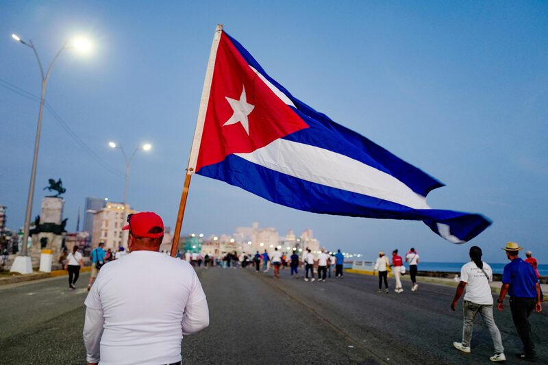 Una persona sostiene una bandera de Cuba mientras camina por el malecón de La Habana, en medio de un grupo de transeúntes. Una persona sostiene una bandera de Cuba mientras camina por el malecón de La Habana, en medio de un grupo de transeúntes.