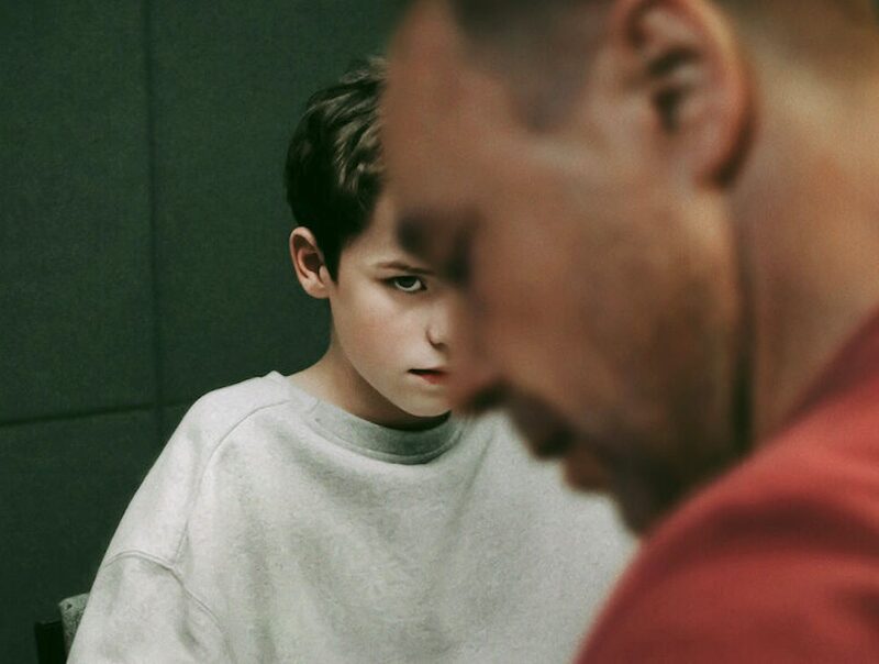 Homem em primeiro plano, desfocado, usando uma camiseta vermelha. Em segundo plano, e em foco, um adolescente com moletom cinza Homem em primeiro plano, desfocado, usando uma camiseta vermelha. Em segundo plano, e em foco, um adolescente com moletom cinza