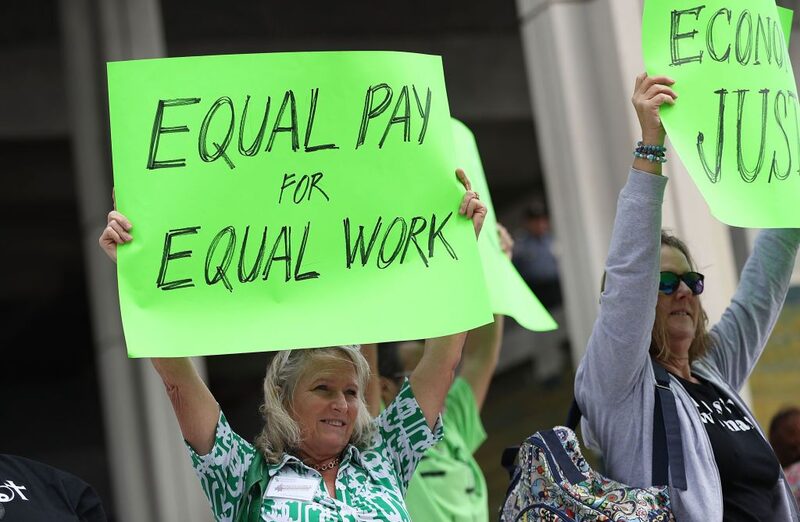 Foto de duas mulheres em protesto. A mulher centralizada carrega um cartaz feito de cartolina verde no qual é possível ler, em inglês, os dizeres "pagamentos iguais por trabalhos iguais" Foto de duas mulheres em protesto. A mulher centralizada carrega um cartaz feito de cartolina verde no qual é possível ler, em inglês, os dizeres "pagamentos iguais por trabalhos iguais"