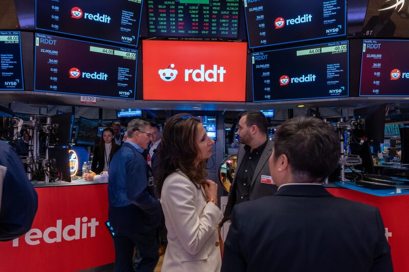 Em primeiro plano, algumas pessoas conversam. Em segundo plano, várias telas de televisão exibindo o logotipo e o código da empresa Reddit Em primeiro plano, algumas pessoas conversam. Em segundo plano, várias telas de televisão exibindo o logotipo e o código da empresa Reddit