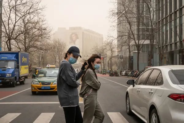 Pedestrians wearing protective masks walk across a road past buildings shrouded in polluted air in Beijing, China, on Monday, March 15, 2021. A sandstorm sweeping across much of northern China left the city in an orange fog and helped push air quality levels in the capital to the worst since 2017.